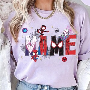 May include: A light purple sweatshirt with the word "NAME" in large, colorful letters. Each letter features a different Spider-Man character and design. Spiderwebs and star accents surround the letters, with a Spider-Man logo on the top left and bottom right.