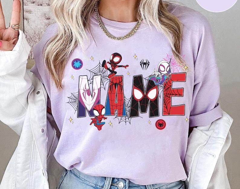May include: A light purple sweatshirt with the word "NAME" in large, colorful letters. Each letter features a different Spider-Man character and design. Spiderwebs and star accents surround the letters, with a Spider-Man logo on the top left and bottom right.
