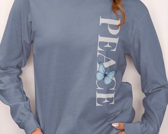Könnte beinhalten: Ein langärmeliges, staubblaues Shirt mit dem Wort „FRIEDEN“ vertikal in Weiß gedruckt. Eine hellblaue Schmetterlingsgrafik befindet sich neben dem Wort „FRIEDEN“. Das Shirt ist aus einem weichen Material gefertigt.