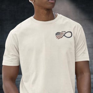 May include: Cream-colored t-shirt featuring a small graphic on the upper left chest. The graphic combines the American flag with an infinity symbol and the year "1776". The shirt has short sleeves and a crew neck.