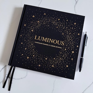 May include: A black journal with the word "LUMINOUS" in gold lettering, surrounded by a constellation design. The journal also has the text "LUNISOLAR PLANNER & GUIDED JOURNAL". A black pen rests on the white marble surface next to the journal.