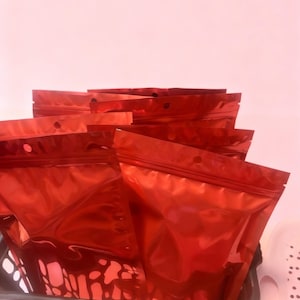 May include: Several shiny red, resealable pouches with a zip closure, presented in a dark grey basket. The pouches have a metallic finish and a small hole near the top. The background is a soft pink.