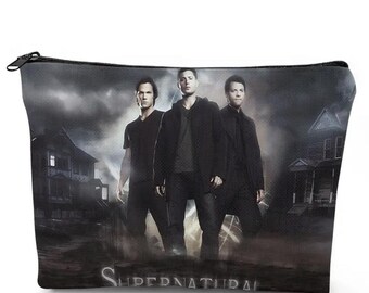 May include: A black zippered cosmetic bag featuring three figures in dark clothing against a stormy background with the word "SUPERNATURAL" at the bottom. The bag has a black zipper and a rectangular shape.