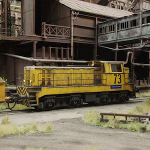 Puede incluir: Una locomotora diésel amarilla, con el número 73, está estacionada en una vía férrea. El fondo muestra edificios industriales e infraestructura. La escena se desarrolla al aire libre, con un ambiente industrial.