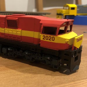 May include: A red and yellow model train locomotive with the text "Tasrail" and the number "2020" printed on the side. The model train has black wheels and a black undercarriage. The train is sitting on a wooden surface.
