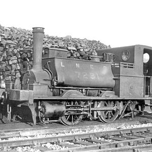 May include: Black and white image of a vintage steam locomotive on a railway track. The locomotive features a cylindrical smokestack, a water tank with the text "LNER 7231", and a small cabin. The background shows a pile of stacked bricks.