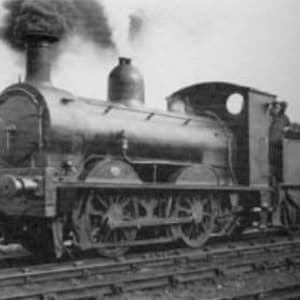 May include: Black and white image of a vintage steam locomotive on tracks. The locomotive has a large smokestack, a rounded front, and multiple wheels. Smoke billows from the stack, suggesting it is in operation. The image is a side view.