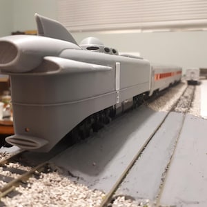 Könnte beinhalten: Ein graues Modell eines Zuges mit einem einzigartigen, stromlinienförmigen Design. Der Zug hat einen markanten vorderen Abschnitt mit einer großen, kreisförmigen Öffnung und einer flossenartigen Struktur oben. Die Waggons sind grau mit roten und weißen Streifen. Das Modell steht auf einem Gleis.