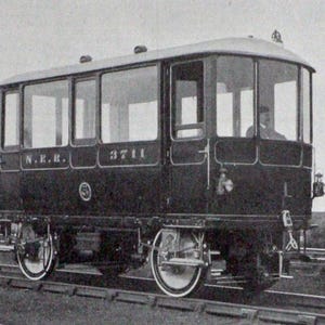 May include: Vintage black and white image of a railway carriage. The carriage has large windows, a rounded roof, and is labeled "N.E.R." and "3711". It is on a railway track.