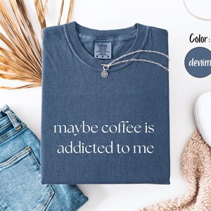 May include: A denim blue t-shirt with the phrase "maybe coffee is addicted to me" printed in white. A silver necklace with a pendant rests on the shirt. The shirt is folded next to a pair of jeans and white shoes.