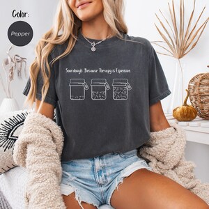 Può includere: T-shirt color pepe con la scritta "Sourdough Because Therapy is Expensive" sopra una grafica di tre barattoli. I barattoli sembrano contenere pasta madre in varie fasi. La maglietta è di vestibilità ampia.