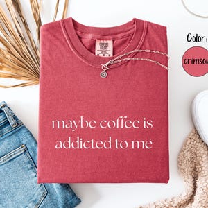 May include: Crimson-colored t-shirt with the phrase "maybe coffee is addicted to me" in white lettering. The shirt is paired with blue jeans, white sneakers, and a silver necklace. A dried palm leaf adds a decorative touch.