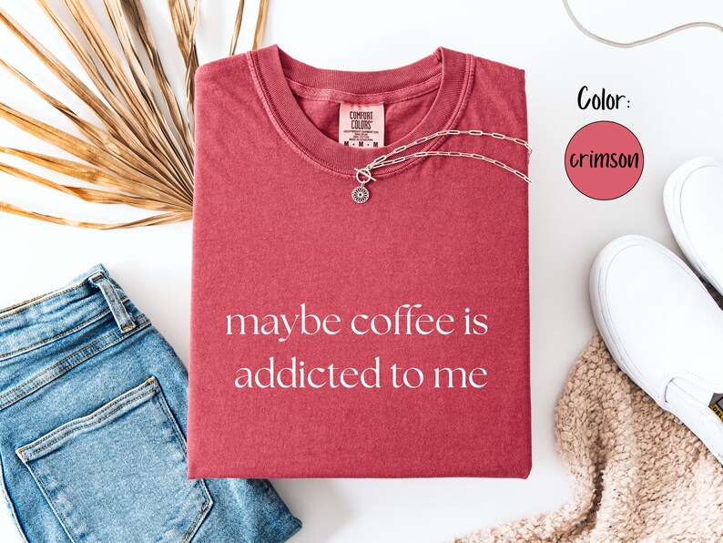 May include: Crimson-colored t-shirt with the phrase "maybe coffee is addicted to me" in white lettering. The shirt is paired with blue jeans, white sneakers, and a silver necklace. A dried palm leaf adds a decorative touch.