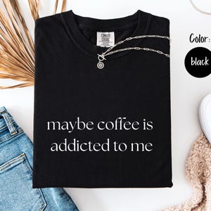 May include: Black t-shirt with the phrase "maybe coffee is addicted to me" in white script. A silver necklace rests on the shirt. The shirt is folded next to blue jeans, white shoes, and a beige sweater.