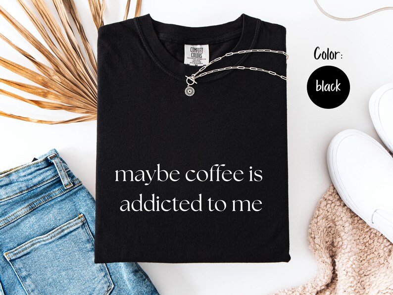 May include: Black t-shirt with the phrase "maybe coffee is addicted to me" in white script. A silver necklace rests on the shirt. The shirt is folded next to blue jeans, white shoes, and a beige sweater.