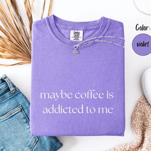 May include: A violet Comfort Colors t-shirt with the phrase "maybe coffee is addicted to me" printed on the front. A silver necklace with a pendant is draped on the shirt. The shirt is paired with blue jeans and white sneakers.