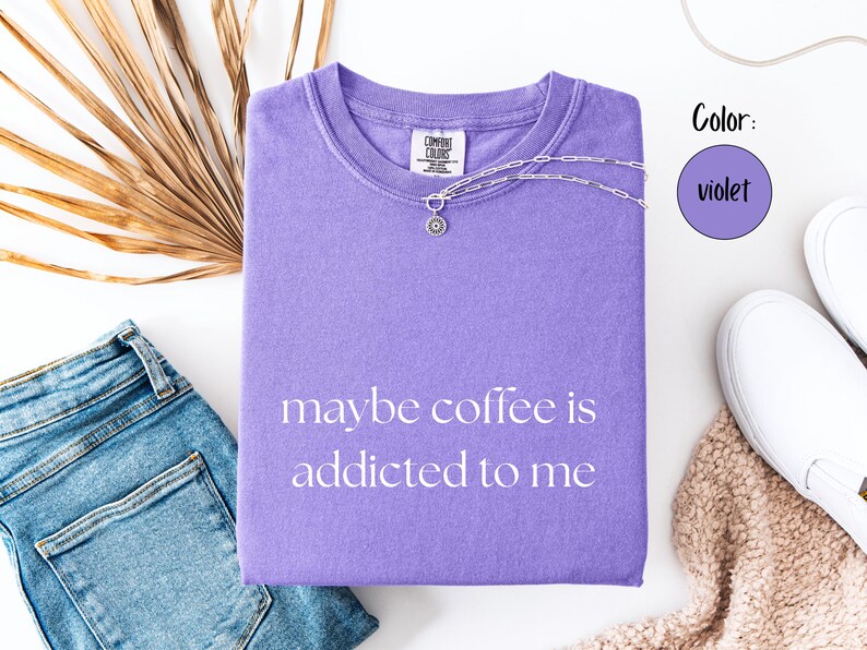 May include: A violet Comfort Colors t-shirt with the phrase "maybe coffee is addicted to me" printed on the front. A silver necklace with a pendant is draped on the shirt. The shirt is paired with blue jeans and white sneakers.
