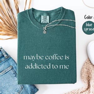 May include: Blue spruce t-shirt with the phrase "maybe coffee is addicted to me" in white. The shirt is folded and accessorized with a silver necklace, blue jeans, and white sneakers. The color is blue spruce.