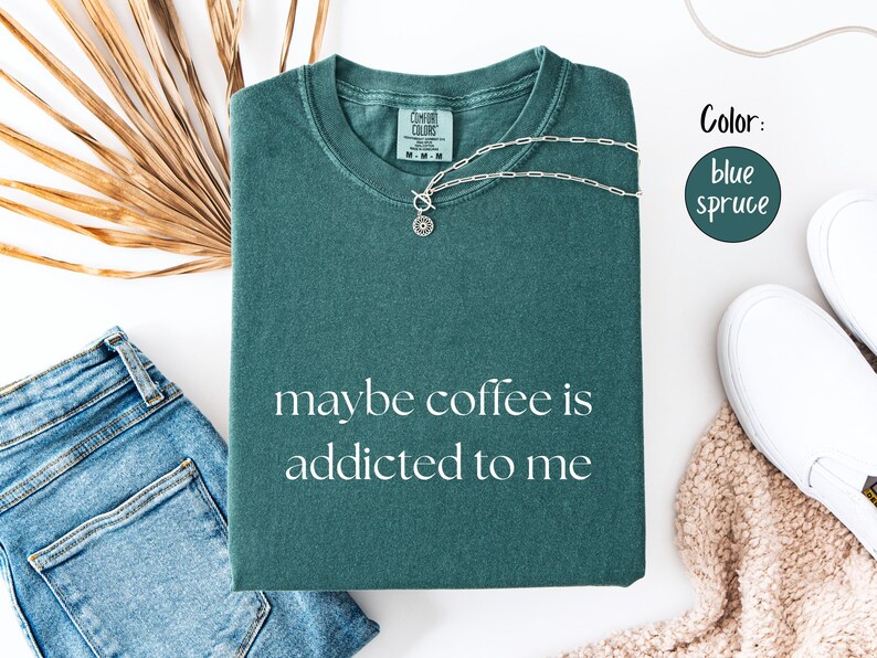 May include: Blue spruce t-shirt with the phrase "maybe coffee is addicted to me" in white. The shirt is folded and accessorized with a silver necklace, blue jeans, and white sneakers. The color is blue spruce.
