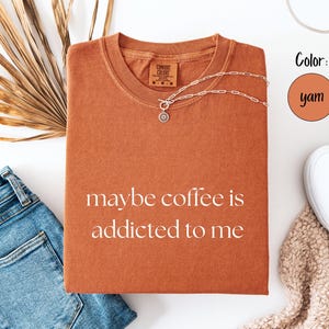 May include: A yam-colored t-shirt with the phrase "maybe coffee is addicted to me" printed in white. A silver necklace with a pendant is on the shirt. The shirt is next to blue jeans, white sneakers, and a dried palm leaf.