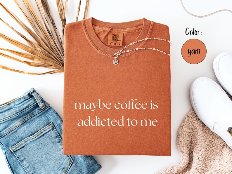 May include: A yam-colored t-shirt with the phrase "maybe coffee is addicted to me" printed in white. A silver necklace with a pendant is on the shirt. The shirt is next to blue jeans, white sneakers, and a dried palm leaf.