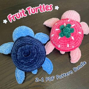 May include: Two plush crochet turtles, one dark blue and one pink strawberry-themed with a green top and white seed accents. The text "Fruit Turtles" and "2-1 PDF Pattern Bundle" is visible in the image, along with small white flower icons.
