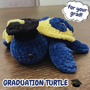 May include: A handmade blue crochet turtle wearing a black graduation cap with yellow trim. The turtle has a yellow and blue shell. Text on the image reads "For your grad!" and "GRADUATION TURTLE".