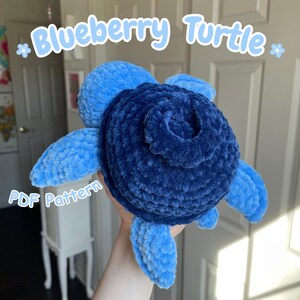 May include: A plush, handmade turtle in shades of blue. The turtle's shell is a deep indigo, resembling a blueberry, with lighter blue flippers and head. The text "Blueberry Turtle" and "PDF Pattern" are visible.