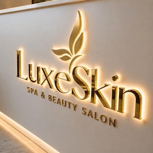 以下が含まれることがあります： LuxeSkin SPA & BEAUTY SALONという金色の文字と、Luxeという単語の上に配置された様式化された葉のアイコンが特徴の立体的な壁面看板。文字はバックライトで照らされており、明るい壁面に柔らかな光を放っています。