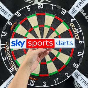 Może przedstawiać: Prostokątna naklejka z napisem sky sports darts w kolorze niebieskim i czerwonym na białym tle, trzymana przed okrągłą czarno-białą tarczą do darta z zielonymi i czerwonymi segmentami punktowymi oraz nadrukowanym słowem Proscore.