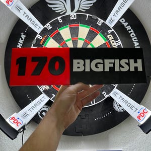 May include: A rectangular sign featuring a split design with a red left side displaying the black text 170 and a black right side displaying the white text BIGFISH. The sign is held in front of a circular dartboard with a black and white frame.