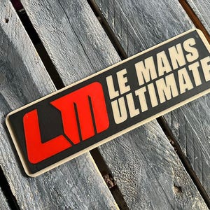 以下が含まれることがあります： 黒い背景に白い文字で「LE MANS ULTIMATE」と書かれた長方形のステッカー。 「LM」の文字は赤色です。 ステッカーはベージュ色の縁取りがあり、風化した木製の表面に置かれています。