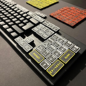 May include: Black mechanical keyboard with custom keycaps. The keycaps feature various symbols and text, including "SORRY", "MUTE", and car-related icons. Additional yellow and red keycap sets are visible in the background.