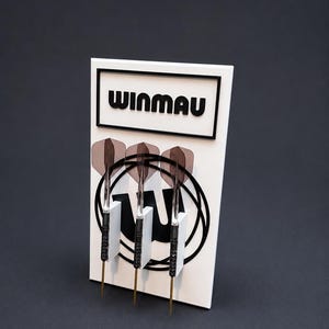 May include: A white rectangular display stand featuring the black text WINMAU at the top. Three darts with black barrels, gold-coloured tips, and translucent dark grey flights are mounted vertically on the front, positioned over a black circular logo.