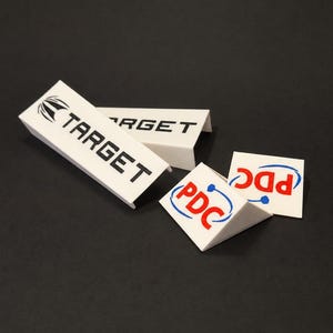 May include: Two white triangular shelf talkers featuring the black text TARGET with a graphic icon, and two white triangular shelf talkers featuring the red text PDC with a blue circular graphic element, all displayed on a dark surface.