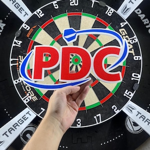 Op de afbeelding: Een driedimensionaal bord met de rode letters PDC met een witte omlijning, geaccentueerd door een blauwe dartflight-afbeelding. Het bord is geplaatst voor een rond dartbord met zwarte, witte, rode en groene segmenten.