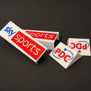 Puede incluir: Marcadores blancos con forma de prisma triangular que presentan el logotipo azul y rojo de Sky Sports y el logotipo rojo y azul de PDC. Estos artículos impresos tienen un diseño angular distintivo con una marca clara en las superficies blancas.