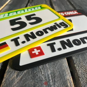 May include: Two racing-style license plates. One is yellow and green with the number 55 and the name T.Norwig. The other is black and white with the same name. Both have flags of Germany and Switzerland.