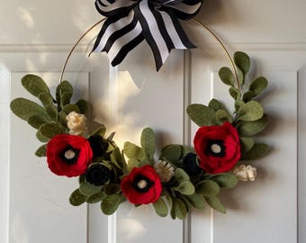 May include: A handmade wreath featuring red felt poppies, green leaves, and black accents on a gold metal ring. A large black and white striped bow is at the top. This decorative wreath is perfect for adding a touch of spring to your home.
