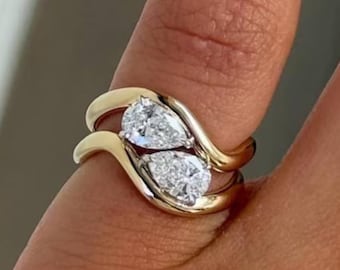 May include: A gold ring featuring two pear-shaped diamonds. The ring has a unique, flowing design with the diamonds set side-by-side. The band is a polished gold color, and the diamonds sparkle brightly.