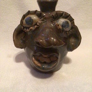 May include: A brown ceramic jug with a face-like design. The jug features large, bulging eyes with blue irises, a prominent nose, and a wide, open mouth. A small, hat-like feature sits atop the jug. The pottery has a rustic, handmade appearance.