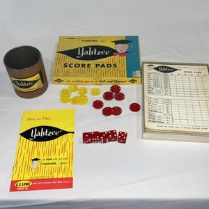 Op de afbeelding: Een vintage Yahtzee-spelset, met een cilindrische container, scorepads, dobbelstenen en fiches. De doos en instructies zijn geel en zwart, met het Yahtzee-logo en speld details. De dobbelstenen zijn rood met witte stippen.