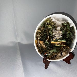 May include: A decorative plate with a landscape scene featuring trees, a stream, and figures. The plate is displayed on a wooden stand. The artwork has a vintage aesthetic with a detailed depiction of nature.
