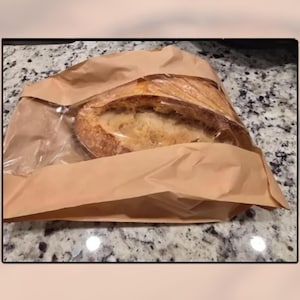 May include: A loaf of crusty bread in a brown paper bag. The bread is partially visible through a clear plastic wrap, showing a golden-brown crust and a soft, airy interior. The bag is open, suggesting the bread is fresh.