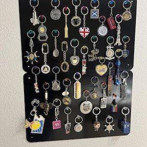May include: A collection of keychains displayed on a black magnetic board. The keychains feature various designs, including landmarks, flags, and novelty items. The keychains are silver, gold, and colorful, with a variety of shapes and sizes.