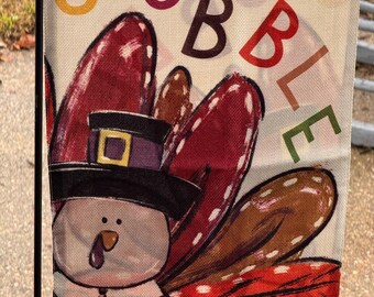 May include: Thanksgiving garden flag featuring a cartoon turkey wearing a pilgrim hat. The flag has the word "GOBBLE" in colorful letters. The turkey has a white face, a red wattle, and a purple and black hat.