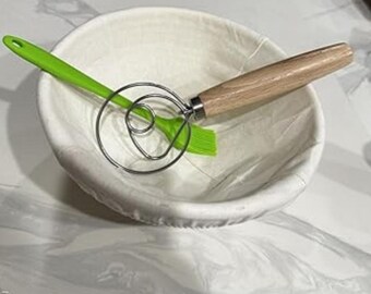 Puede incluir: Una cesta de fermentación redonda blanca con un cepillo de silicona verde, un batidor de masa de acero inoxidable y un batidor de masa con mango de madera. Las herramientas son para hornear pan.