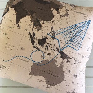 Op de afbeelding: Een beige kussen met een wereldkaart erop gedrukt. De kaart is bruin en zwart, met een blauw papieren vliegtuig dat over Azi&euml; vliegt en een blauwe stippellijn die Australi&euml; omcirkelt. De tekst "Indische Oceaan" en "Australi&euml;" is zichtbaar op de kaart.