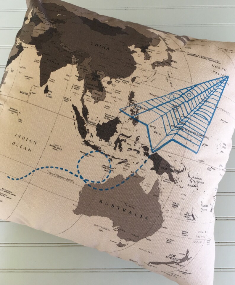 Op de afbeelding: Een beige kussen met een wereldkaart erop gedrukt. De kaart is bruin en zwart, met een blauw papieren vliegtuig dat over Azi&euml; vliegt en een blauwe stippellijn die Australi&euml; omcirkelt. De tekst "Indische Oceaan" en "Australi&euml;" is zichtbaar op de kaart.