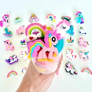 May include: A collection of colorful unicorn and rainbow charms and decorations. A clear container is filled with layers of pink, yellow, and blue sand. A large pink unicorn with a rainbow mane and tail is attached to the container.